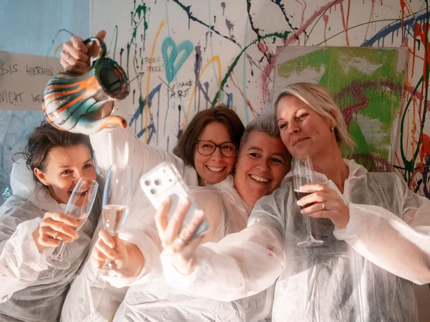 251008_Imagefotos_SplashBox-Einbeck_049-001.JPG Vier Frauen in Schutzanzügen mit Sekt vor bunter Farbspritzwand beim Selfie.