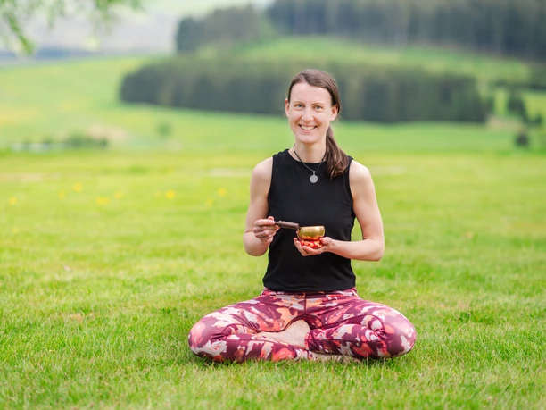 Roots an Flow Frau im Yogastil sitzt im Gras, hält eine Klangschale und lächelt. Grüne Wiesen im Hintergrund.