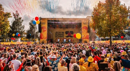 Vor einer Open-Air-Bühne, die karnevalistisch dekoriert ist, steht ein bunt kostümiertes Publikum.