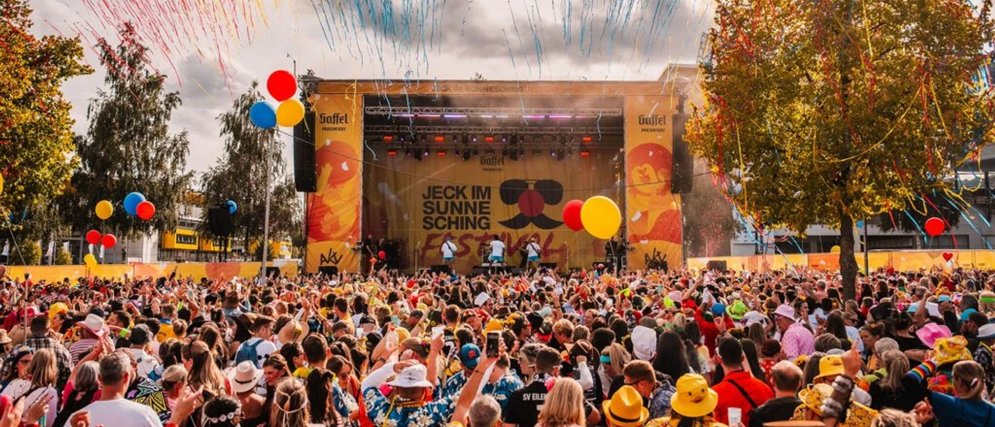 Vor einer Open-Air-Bühne, die karnevalistisch dekoriert ist, steht ein bunt kostümiertes Publikum.