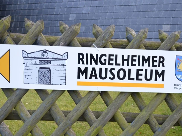 Hinweisschild zum Ringelheimer Mausoleum mit Pfeil, Gebäudeskizze und Wappen an Holzzaun befestigt