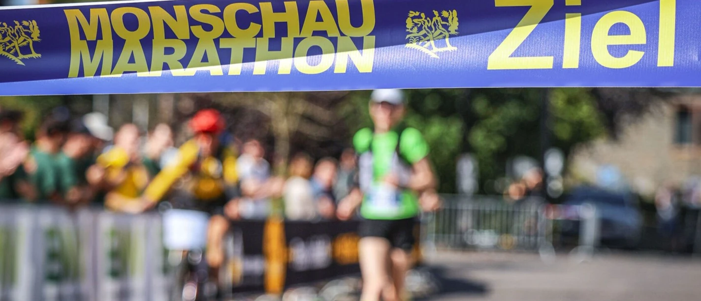 Im Vordergrund ist das Zielband des Monschauer Marathons aufgespannt. Eine Läuferin läuft auf einer abgesperrten Strecke mit Zuschauern darauf zu.