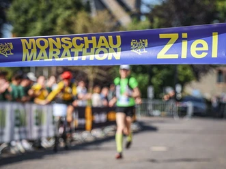 Monschau Marathon (c) Monschau Marathon.jpg Im Vordergrund ist das Zielband des Monschauer Marathons aufgespannt. Eine Läuferin läuft auf einer abgesperrten Strecke mit Zuschauern darauf zu.
