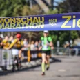 Monschau Marathon (c) Monschau Marathon.jpg Im Vordergrund ist das Zielband des Monschauer Marathons aufgespannt. Eine Läuferin läuft auf einer abgesperrten Strecke mit Zuschauern darauf zu.