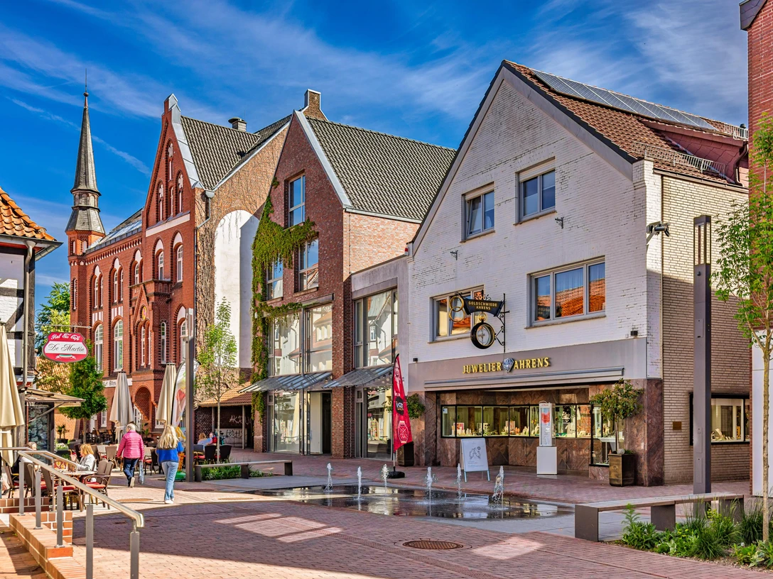 1208_2025_FotografieDiepholz.jpg Belebte Einkaufsstraße in Diepholz mit historischen Gebäuden, kleinen Geschäften und Wasserspielen.
