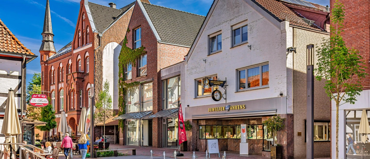 1208_2025_FotografieDiepholz.jpg Belebte Einkaufsstraße in Diepholz mit historischen Gebäuden, kleinen Geschäften und Wasserspielen.