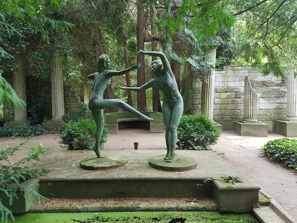 20200707_114233_bremerhaven_erlebnis_Thieles Garten (c) Tanja Albert.jpg Bronzefiguren tanzender Frauen vor alten Steinsäulen, umgeben von dichtem Grün in Thieles Garten.