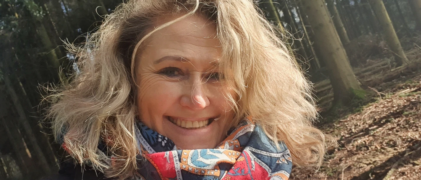 Sabine Fuchs Sabine Fuchs lächelt im sonnigen Wald, mit blondem Haar und buntem Schal.