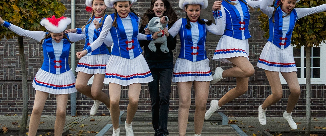 Büttenabend 2026.2.png Sechs Mädchen tanzen in blauen Kostümen. Eine Person hält ein Stofftier. Alle springen fröhlich.
