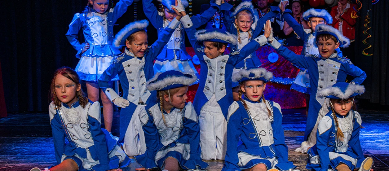 Kinderkarneval 2026.2png.png Kinder tanzen auf einer Bühne. Sie tragen blaue Kostüme mit weißen Hüten.