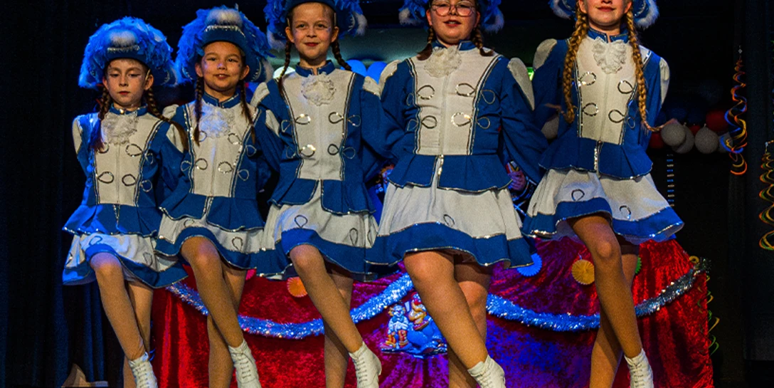 Senioren Nachmittag 2026.2png.png Fünf Kinder tanzen auf einer Bühne. Sie tragen blaue Uniformen und weiße Hüte.