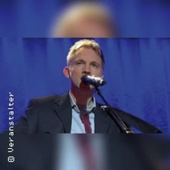 Ein Mann im Anzug steht vor blauem Vorhang und singt in ein Mikrofon.