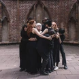 Isaac Chong Wai, Die Mütter, 2022 Gruppe in schwarzer Kleidung umarmt sich eng vor einer Backsteinwand mit gotischen Bögen.Group in black clothing hugging each other in front of a brick wall with Gothic arches.