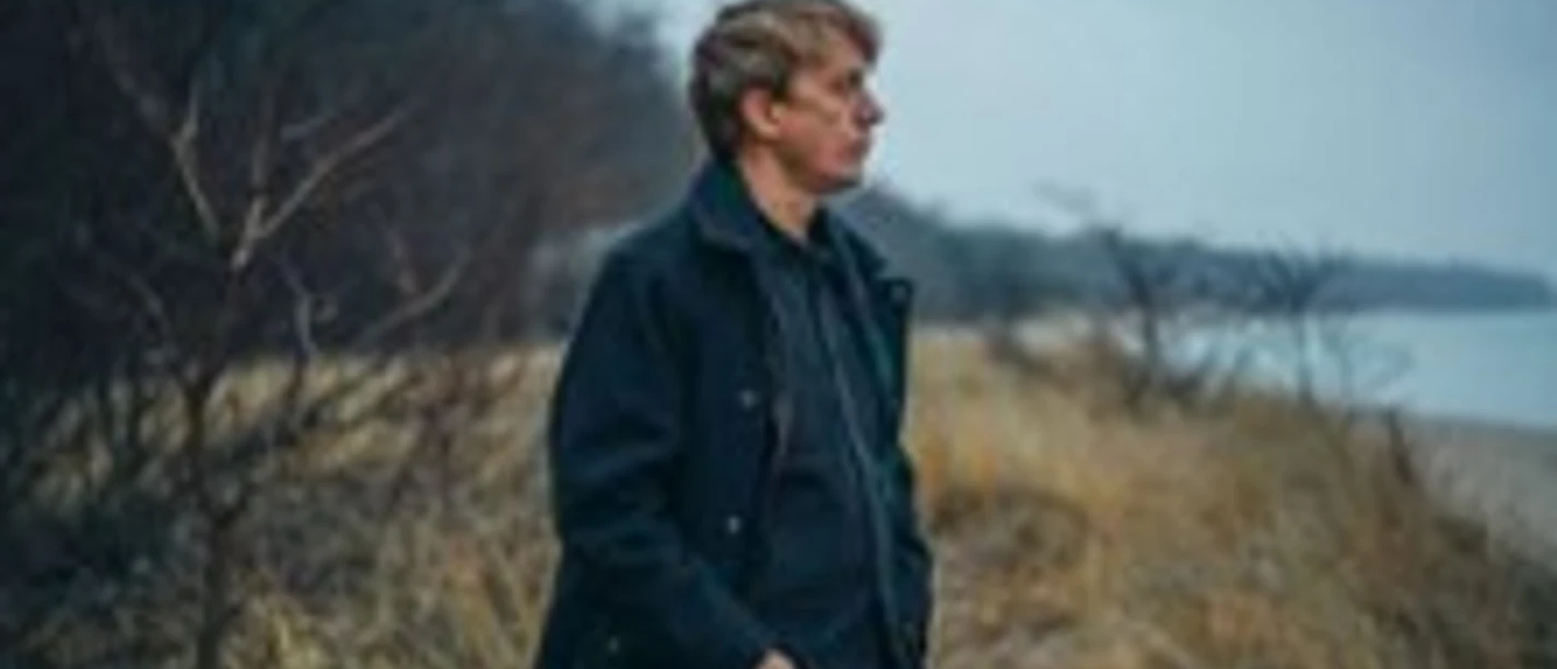 Eine Person steht am Strand neben trockenem Gras unter grauem Himmel und blickt zur Seite.