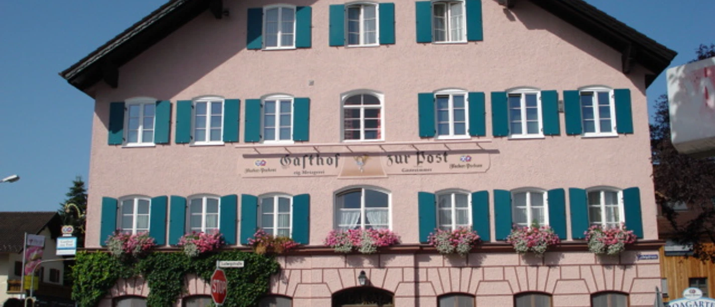 Gasthof zur Post.jpg Großes rosa Haus mit Blumen an Fenstern. Blaue Läden geben freundliche Wirkung.