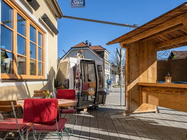 Gasthof Sonne Sonniger Außenplatz mit Tischen, roten Stühlen und einer alten Gondel am Weg.
