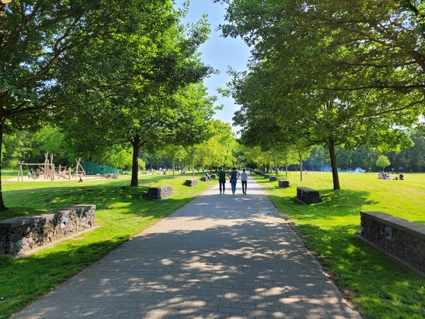 20230604_161253 (c) Tanja Mehl_Erlebnis Bremerhaven.jpg Breiter Weg durch grünen Park mit Schatten spendenden Bäumen, Spielplatz und spazierenden Menschen.
