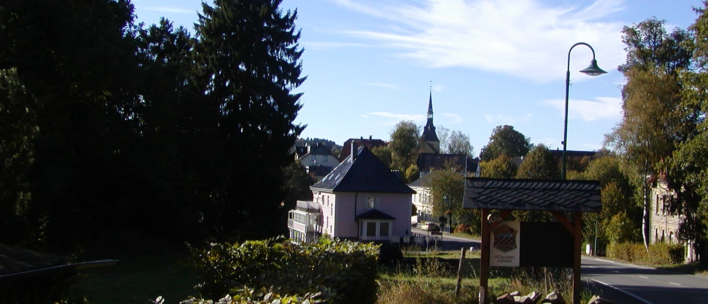 Ortseingang Rönsahl Im Vordergrund links Hecke, Wiese und Bäume. Rechts die Straße nach Rönsahl. Willkommenstafel am Straßenrand. Im Hintergrund Häuser. Am Horizont blauer Himmel mit weißer Wolke und die Turmspitze der Servatiuskirche.