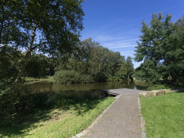 Erikasee M9036 (c) Helmut Gross_Erlebnis Bremerhaven.jpg Schmaler Weg führt zu einem Holzsteg am ruhigen Erikasee, umgeben von Bäumen unter blauem Himmel.