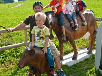 Schönegger Käse-Alm Kinder sitzen auf Kuh-Figuren auf einer Wiese und lachen fröhlich.