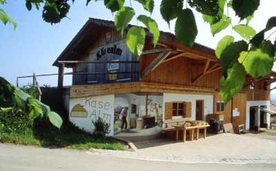 Schönegger Käse-Alm Holzhaus mit Käse-Alm Bild, Sitzbank und grünen Blättern im Vordergrund.