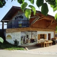 Schönegger Käse-Alm Holzhaus mit Käse-Alm Bild, Sitzbank und grünen Blättern im Vordergrund.