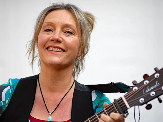 Presefoto von Bettina Göschl Frau mit Gitarre lächelt in die Kamera und trägt eine bunte Bluse sowie eine Halskette mit Anhänger.