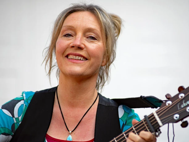 Presefoto von Bettina Göschl Frau mit Gitarre lächelt in die Kamera und trägt eine bunte Bluse sowie eine Halskette mit Anhänger.