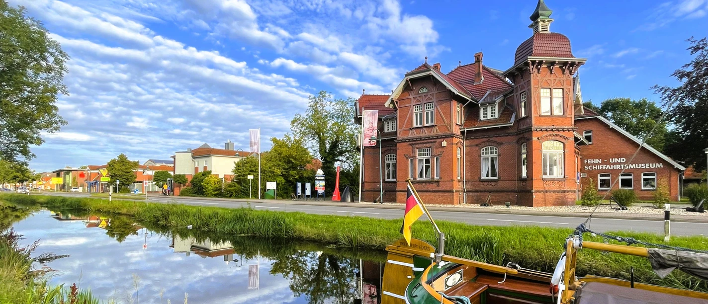 Fehn-und Schiffahrtsmuseum Westrhauderfehn mit Schäfchenwolken am Himmel und davor der Rajen mit Schiffsbug des Plattbodenschiffes Twe.png Das Fehn- und Schiffahrtsmuseum in Westrhauderfehn mit einem grünen Plattbodenschiff im Vordergrund auf dem Wasser und Schäfchenwolken am blauen Himmel.