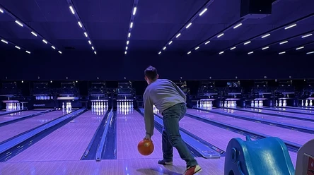 Person beim Bowling in beleuchteter Halle, wirft orange Kugel auf leuchtende Pins unter blauem Licht.