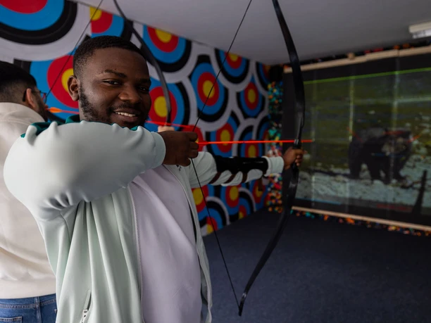 Bogensport Olymp-072 (c) Drei eins Kommunikation GmbH, Christoph Schmitz_Erlebnis Bremerhaven.jpg Ein Mann spannt konzentriert einen Bogen in einem Bogenschießkino mit digitaler Zielwand.