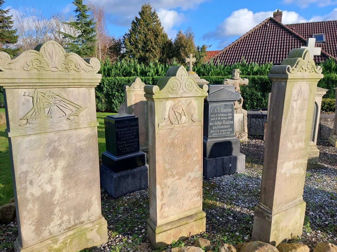 20230117_135003.jpg Drei verwitterte Sandstein-Gedenksteine mit Reliefs und Inschriften auf einem Friedhof, im Hintergrund Hecke und Haus mit rotem Ziegeldach.