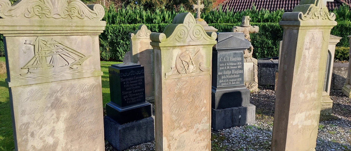 20230117_135003.jpg Drei verwitterte Sandstein-Gedenksteine mit Reliefs und Inschriften auf einem Friedhof, im Hintergrund Hecke und Haus mit rotem Ziegeldach.