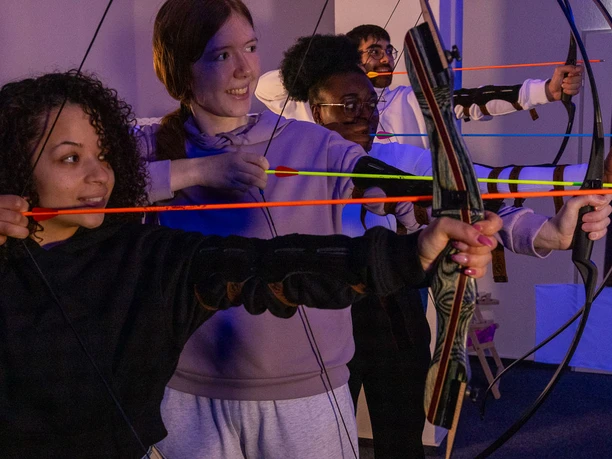 Bogensport Olymp-004 (c) Drei eins Kommunikation GmbH, Christoph Schmitz_Erlebnis Bremerhaven.jpg Vier Personen spannen ihre Bögen im Indoor-Bogenschießkino und zielen konzentriert auf die Zielwand.