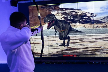 Bogensport Olymp-001 (c) Drei eins Kommunikation GmbH, Christoph Schmitz_Erlebnis Bremerhaven.jpg Person zielt mit Pfeil und Bogen auf Dinosaurier-Projektion im Bogenschießkino in Bremerhaven.
