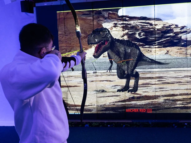 Bogensport Olymp-001 (c) Drei eins Kommunikation GmbH, Christoph Schmitz_Erlebnis Bremerhaven.jpg Person zielt mit Pfeil und Bogen auf Dinosaurier-Projektion im Bogenschießkino in Bremerhaven.