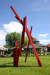 Skulptur, Großer Kranich Rote Metallskulptur mit kantigen, aufragenden Formen auf Rasenfläche vor Gebäuden im Freien.