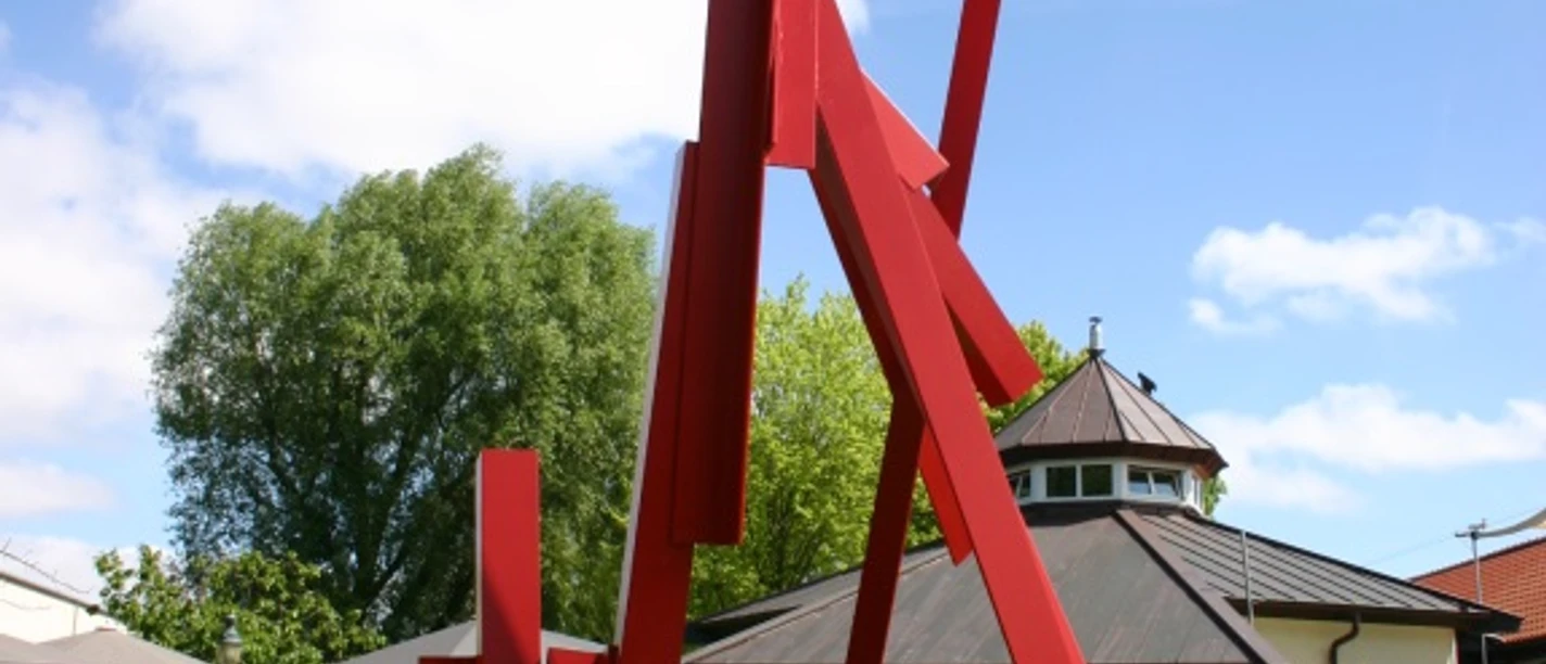 Skulptur, Großer Kranich Rote Metallskulptur mit kantigen, aufragenden Formen auf Rasenfläche vor Gebäuden im Freien.