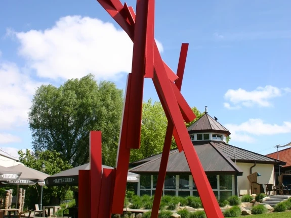 Skulptur, Großer Kranich Rote Metallskulptur mit kantigen, aufragenden Formen auf Rasenfläche vor Gebäuden im Freien.