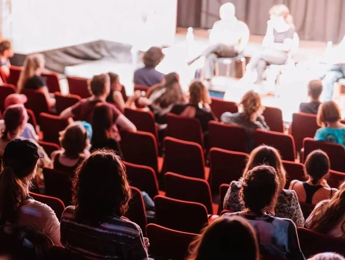 Festival@Annika Bethan Publikum sitzt in roten Theatersesseln und verfolgt eine beleuchtete Bühnenrunde.The audience sits in red theater seats and follows an illuminated stage round.