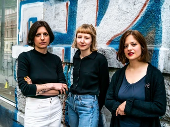 kollektiv flexen Drei Personen stehen vor einer bunt besprühten Wand und blicken ruhig in die KameraThree people stand in front of a colorfully spray-painted wall and look calmly into the camera