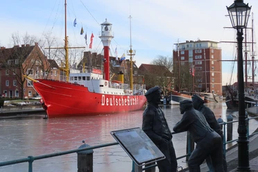 Feuerschiff-Deutsche Bucht.jpg Rotes Feuerschiff mit der Aufschrift »Deutsche Bucht« im Hafen, davor zwei bronzene Figuren an einem Geländer und eine Laterne.