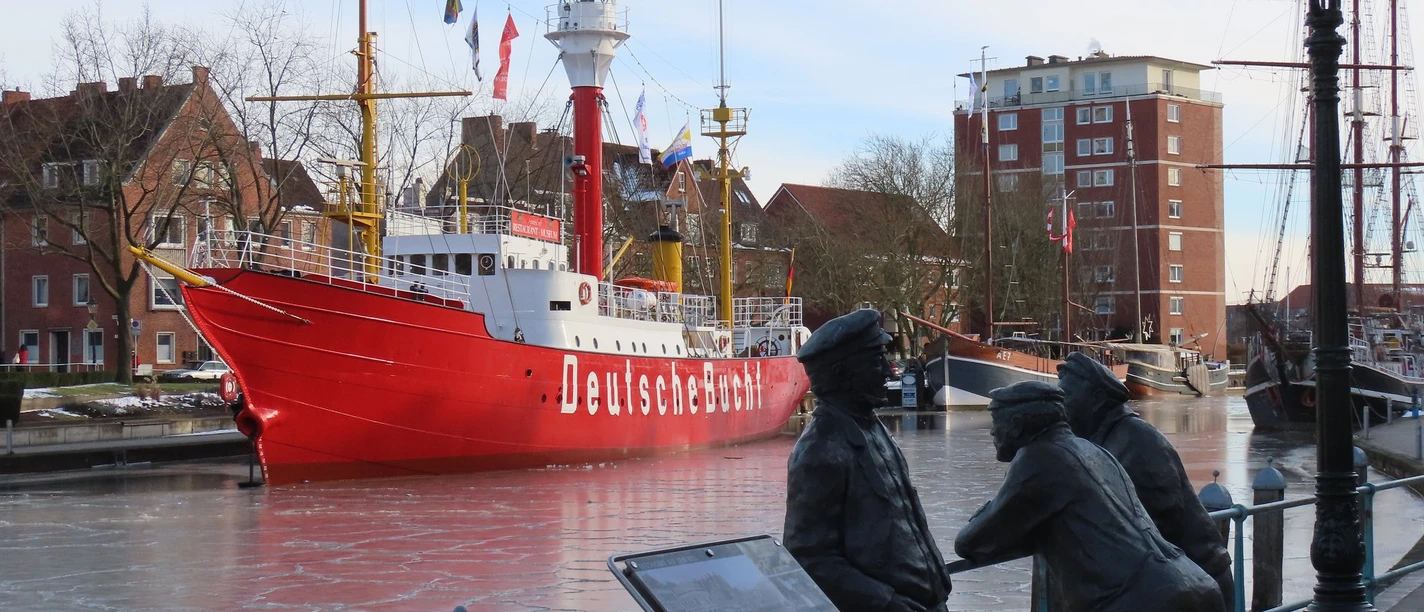 Feuerschiff-Deutsche Bucht.jpg Rotes Feuerschiff mit der Aufschrift »Deutsche Bucht« im Hafen, davor zwei bronzene Figuren an einem Geländer und eine Laterne.