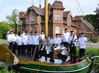 Shanty-Chor Overledinger Jungs Collinghorst.jpg Gruppe von Männern in weißen Matrosenhemden auf einem historischen Segelboot vor einem Backsteingebäude mit der Aufschrift 'FEHN- UND SCHIFFFAHRTSMUSEUM'