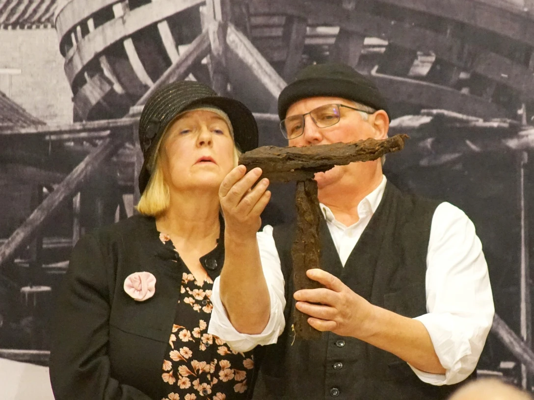 FehnTheater.jpg Zwei Personen vor einem historischen Hintergrund halten ein großes, rostiges Kreuz aus Metall