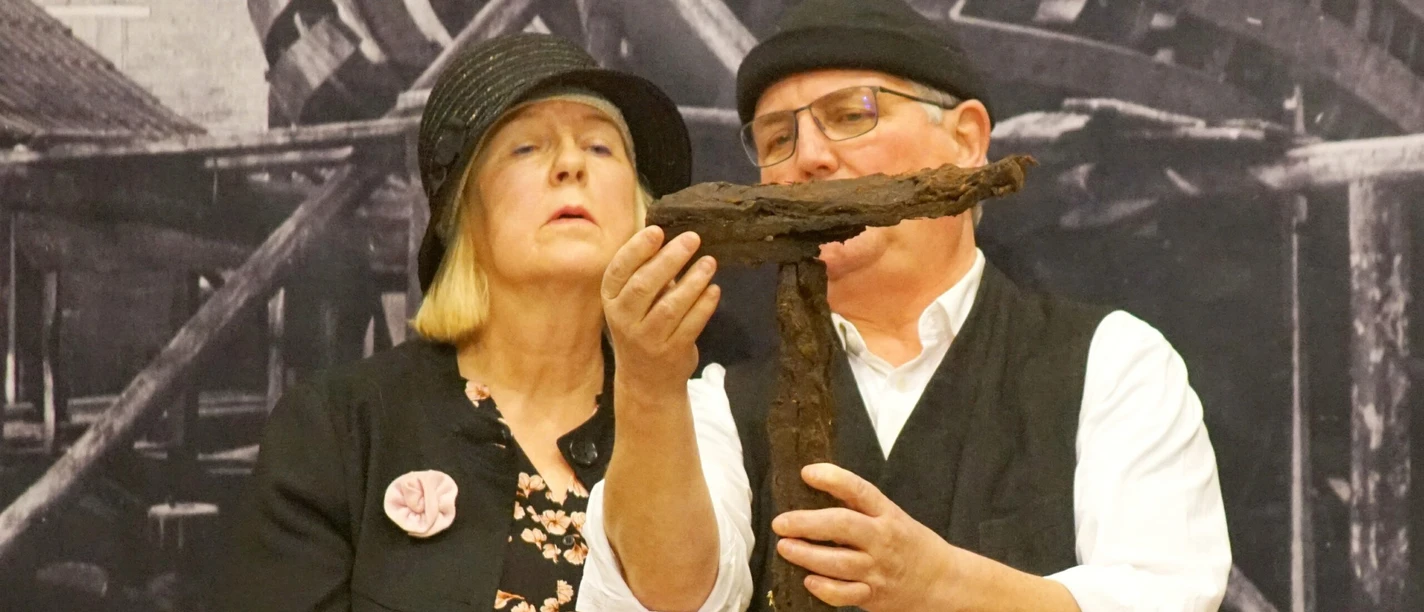 FehnTheater.jpg Zwei Personen vor einem historischen Hintergrund halten ein großes, rostiges Kreuz aus Metall