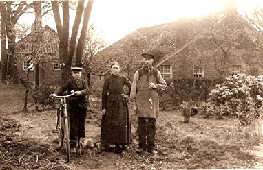 Scannen0029.jpg Drei Personen in historischer Kleidung stehen vor einem Reetdachhaus, eine Person hält ein Fahrrad, eine kleine Kuh ist zu ihren Füßen.