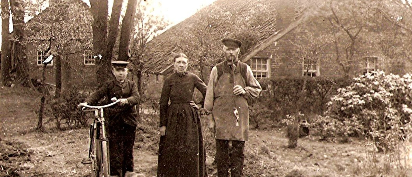 Scannen0029.jpg Drei Personen in historischer Kleidung stehen vor einem Reetdachhaus, eine Person hält ein Fahrrad, eine kleine Kuh ist zu ihren Füßen.