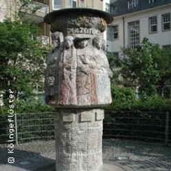 Steinsäule mit figürlichen Reliefs im Außenbereich, umgeben von Bäumen und Gebäuden