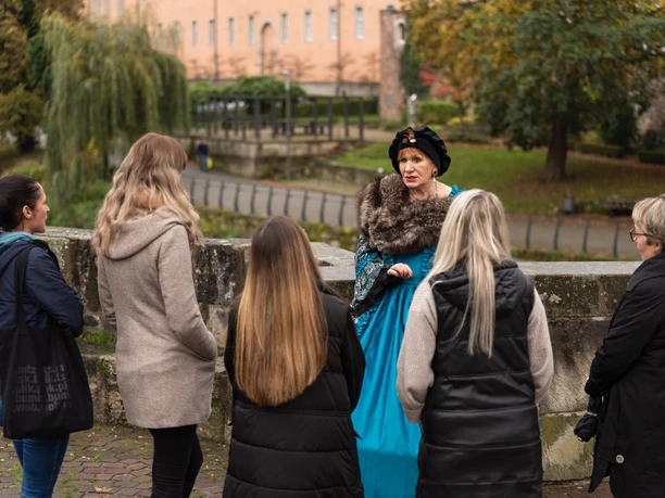 Stadtführung - Hann. Münden Marketing GmbH - Jules Stolzenhain-54.jpg Eine Frau in historischem Kostüm führt eine Gruppe auf einer Stadtführung im Freien an.